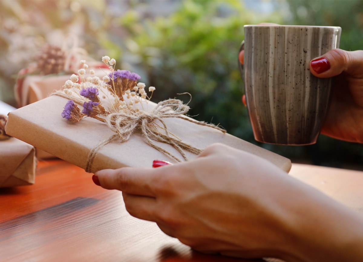 A hand holding a book wrapped in brown gift wrap, tied up with string and small purple and white flowers attached to it.