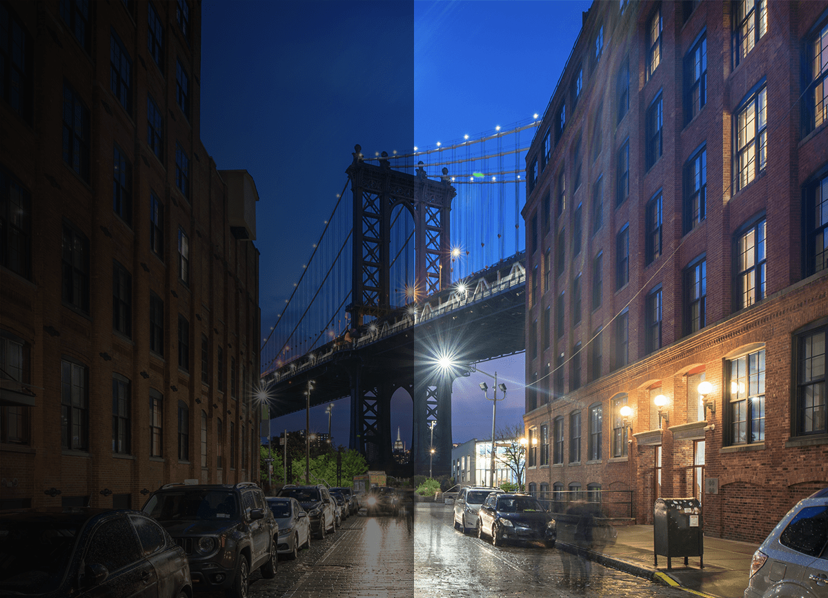 Een afbeelding van een straat 's nachts met de Brooklyn Bridge aan het einde van de straat. De helft van het beeld is donker en nauwelijks zichtbaar, de andere helft is helderder en de mensen erin zijn wazig.