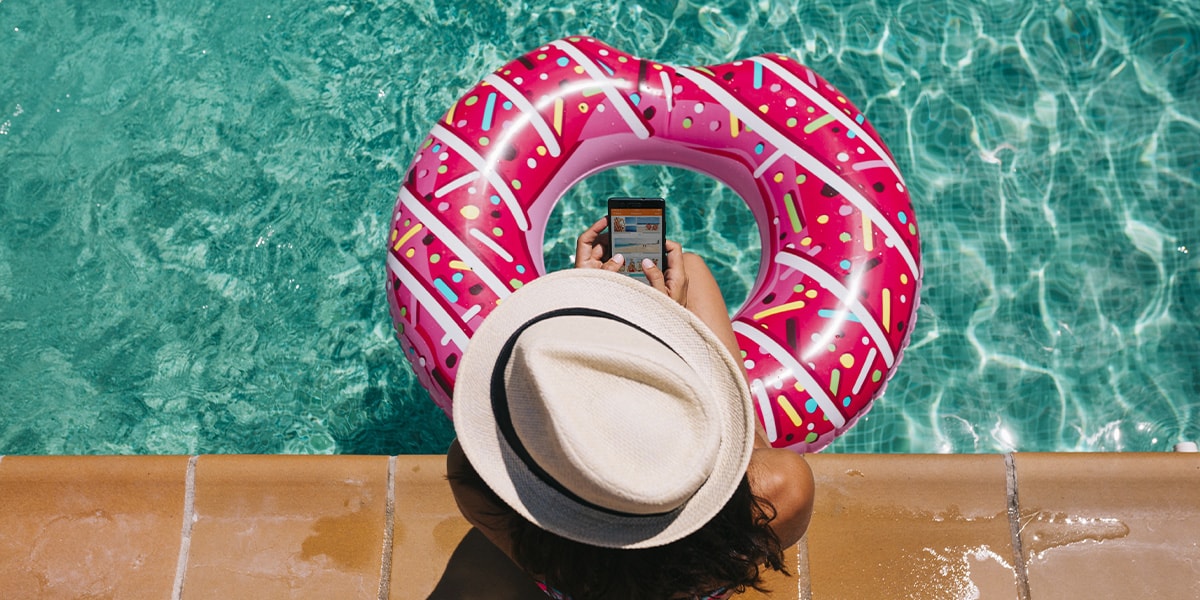 En kvinna sitter vid poolen med benen i en rosa gummiring och håller i en mobil.