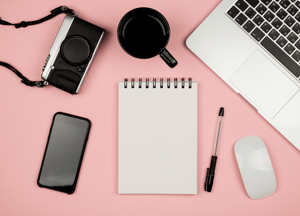 A topdown photo of a laptop, camera, notepad, pen, phone, computer mouse and a mug on a pale pink surface.