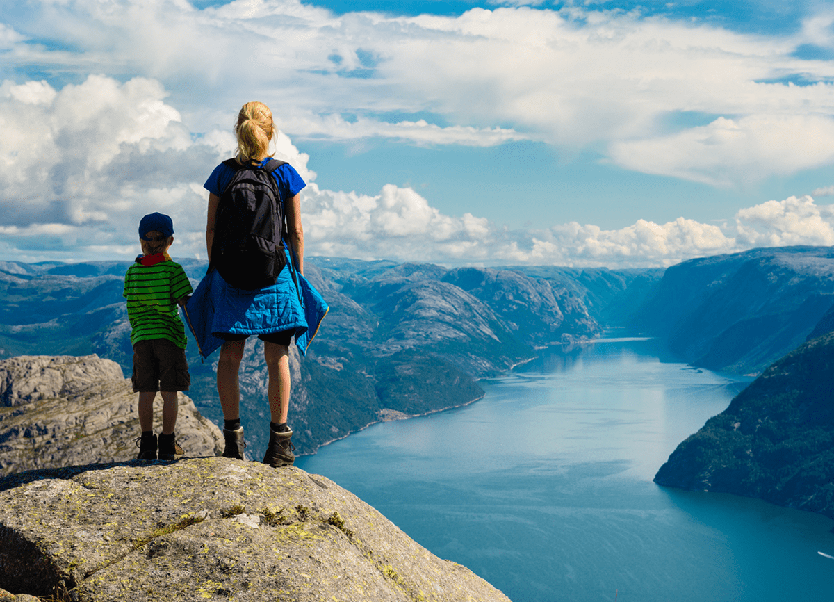 A woman and a young boy in hiking gear standing on a hill top with their backs to the camera, overlooking a river surrounded by mountains.