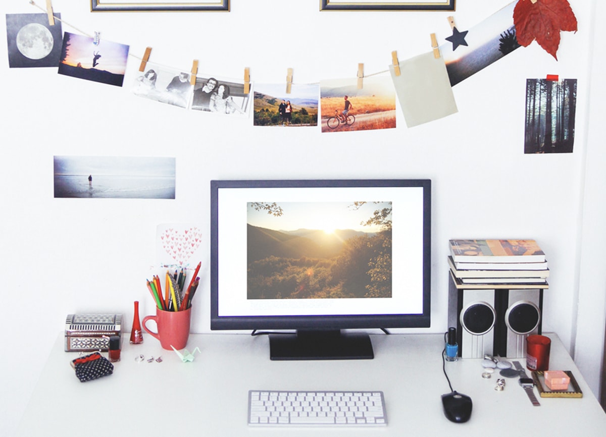 A photo of a white desk in a white room, with a computer, speakers, keyboard, mouse, stationery and books on the desk, and a selection of photos clipped to a string hanging above it.