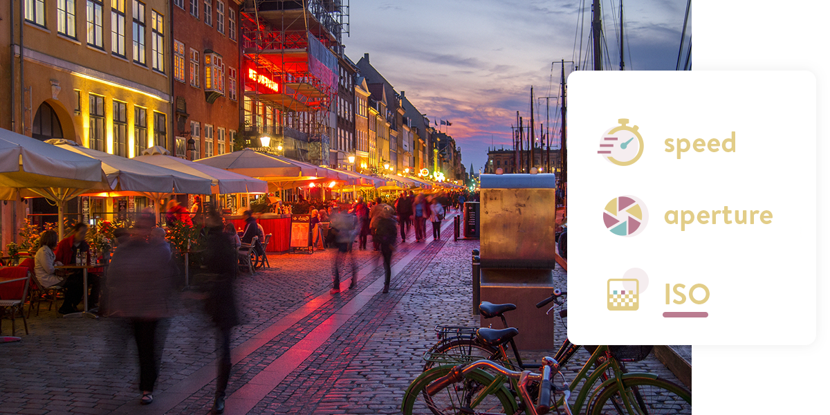 A photo of a busy cobbled street at dusk. The people walking down the street are slightly blurred