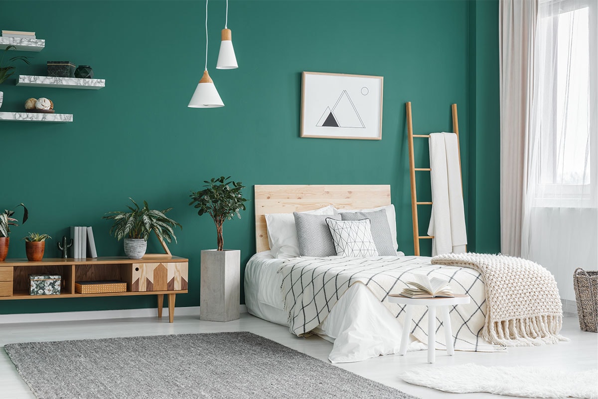 A bedroom with an emerald green feature wall, with a bed covered with white, grey, and tan bedding, marble shelves on the wall and a wooden side table.