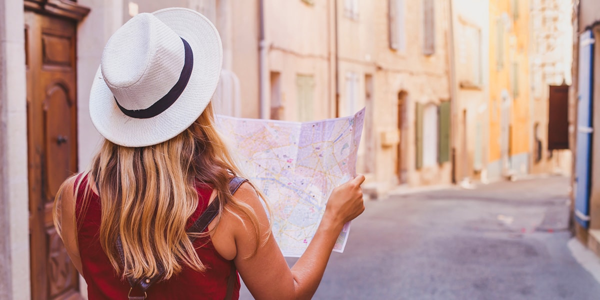 A woman on a city break, with her back facing the camera, looking at a map in a quiet street on a sunny day.