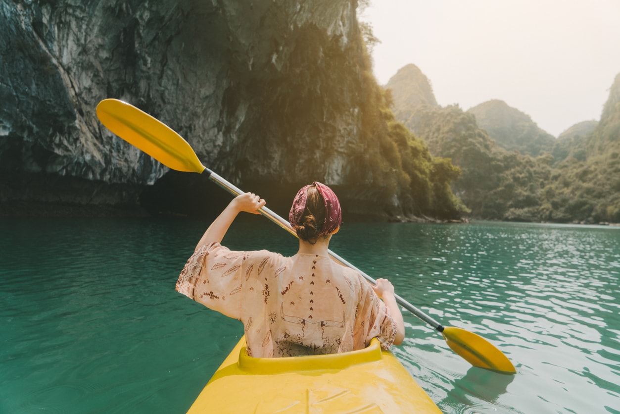 A girl with her back facing the camera, kayaking on the sea. There are mountains and cliffs in the background that are slightly blurred.