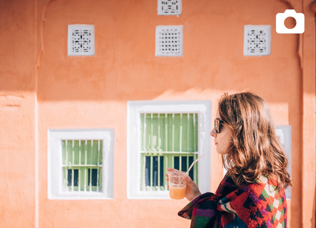 A girl stood outside against a bright orange wall, drinking an orange juice.