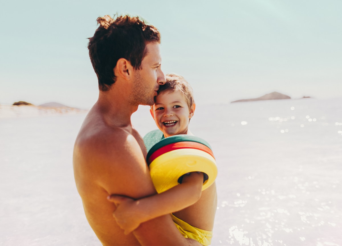 A man holding a little kid on a very bright sunny day at the beach, standing on the shore.