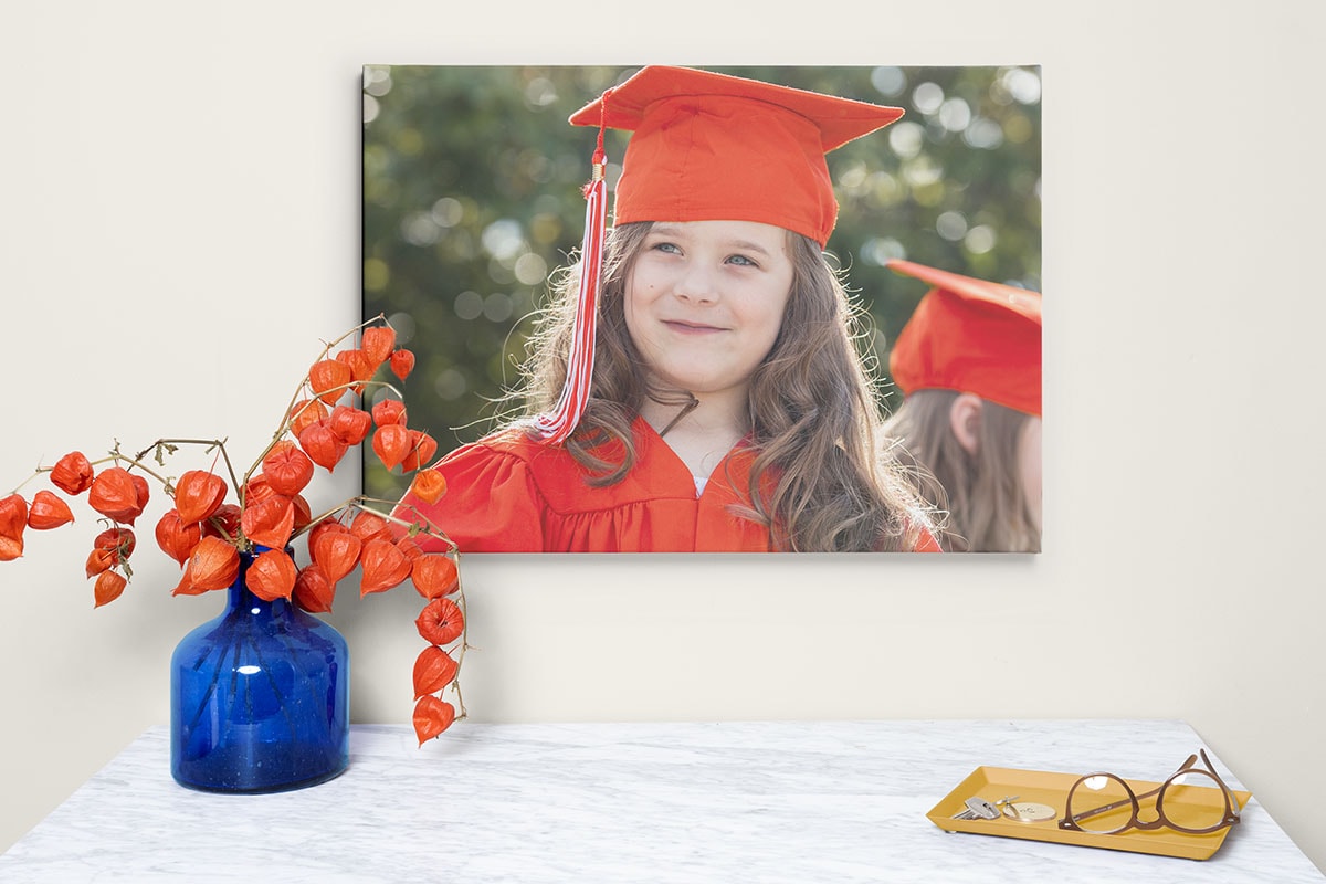 A photo of a framed canvas wall art with a girl’s graduation photo on. It’s hanging on a wall next to a side table with a vase on.