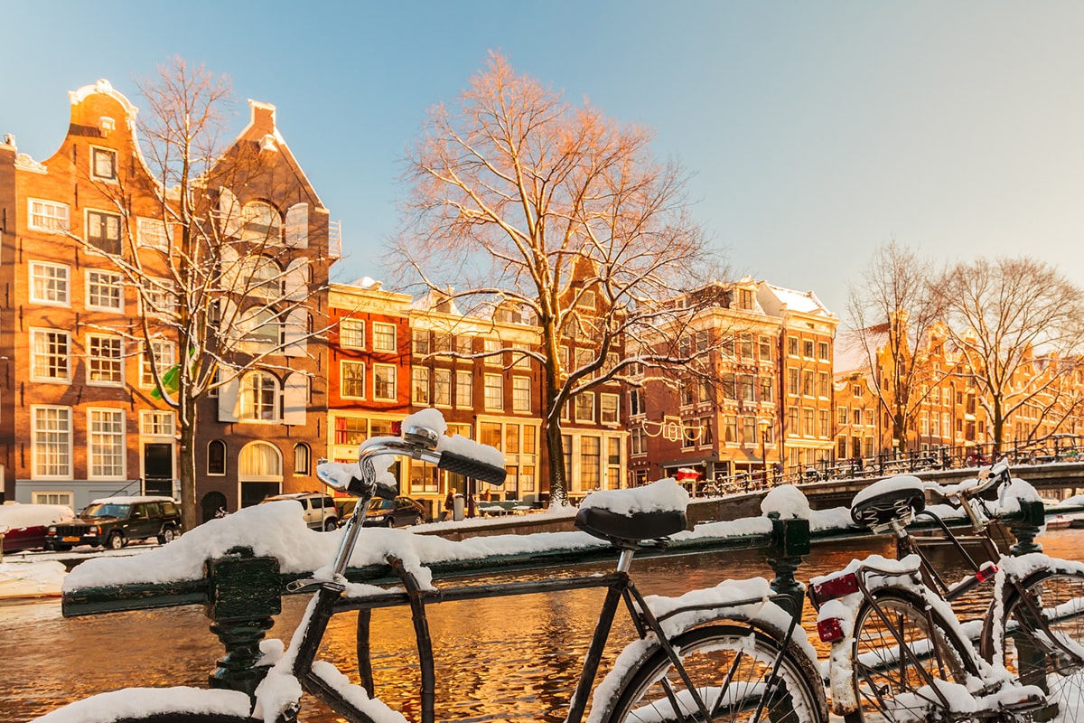 Ein Bild von Amsterdam an einem hellen Wintertag mit einer Reihe schneebedeckter Fahrräder neben einer Gracht.