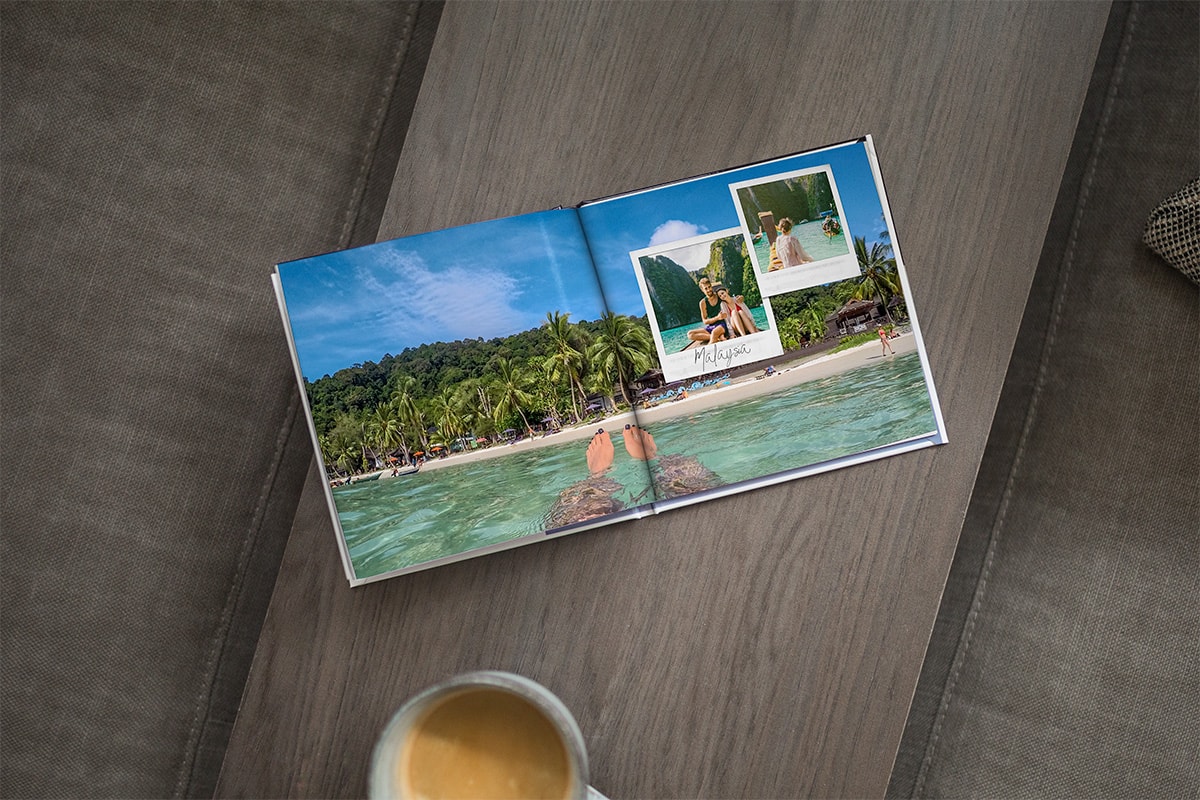 Ein aufgeschlagenes Fotobuch auf einer Holztischplatte. Man sieht ein doppelseitiges Bild einer Frau, die im Meer treibt, mit einem Strand und Palmen im Hintergrund und zwei Fotos im Stil einer Sofortbildkamera in der oberen rechten Ecke.