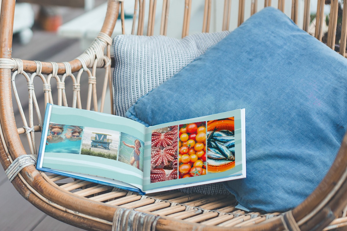 Ein aufgeschlagenes Sommerfotobuch, das auf einem Korbstuhl mit blauen Kissen liegt. Auf der linken Seite befinden sich Sommerurlaubsfotos und auf der rechten Seite sommerliche Fotos von Essen.