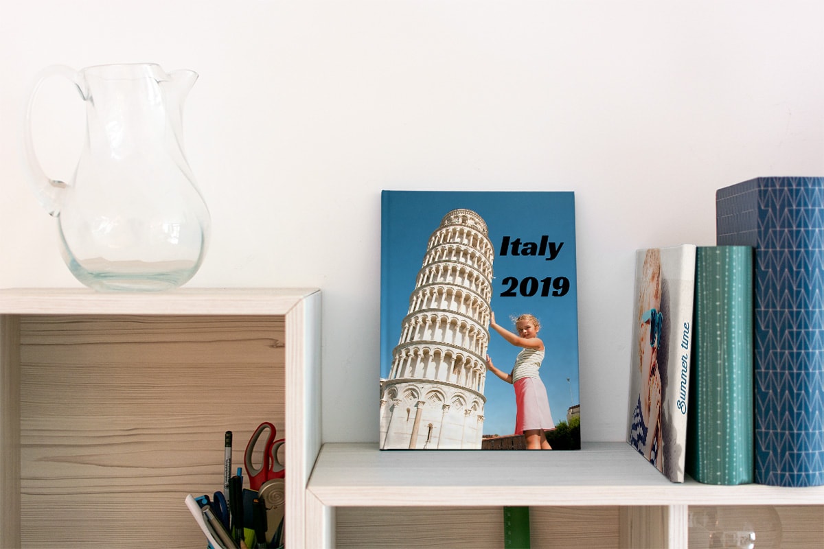Ein Sommerfotobuch auf einem Bücherregal, mit einem kleinen Mädchen, das auf dem Einband posiert und so tut, als würde sie den Schiefen Turm von Pisa umstoßen.