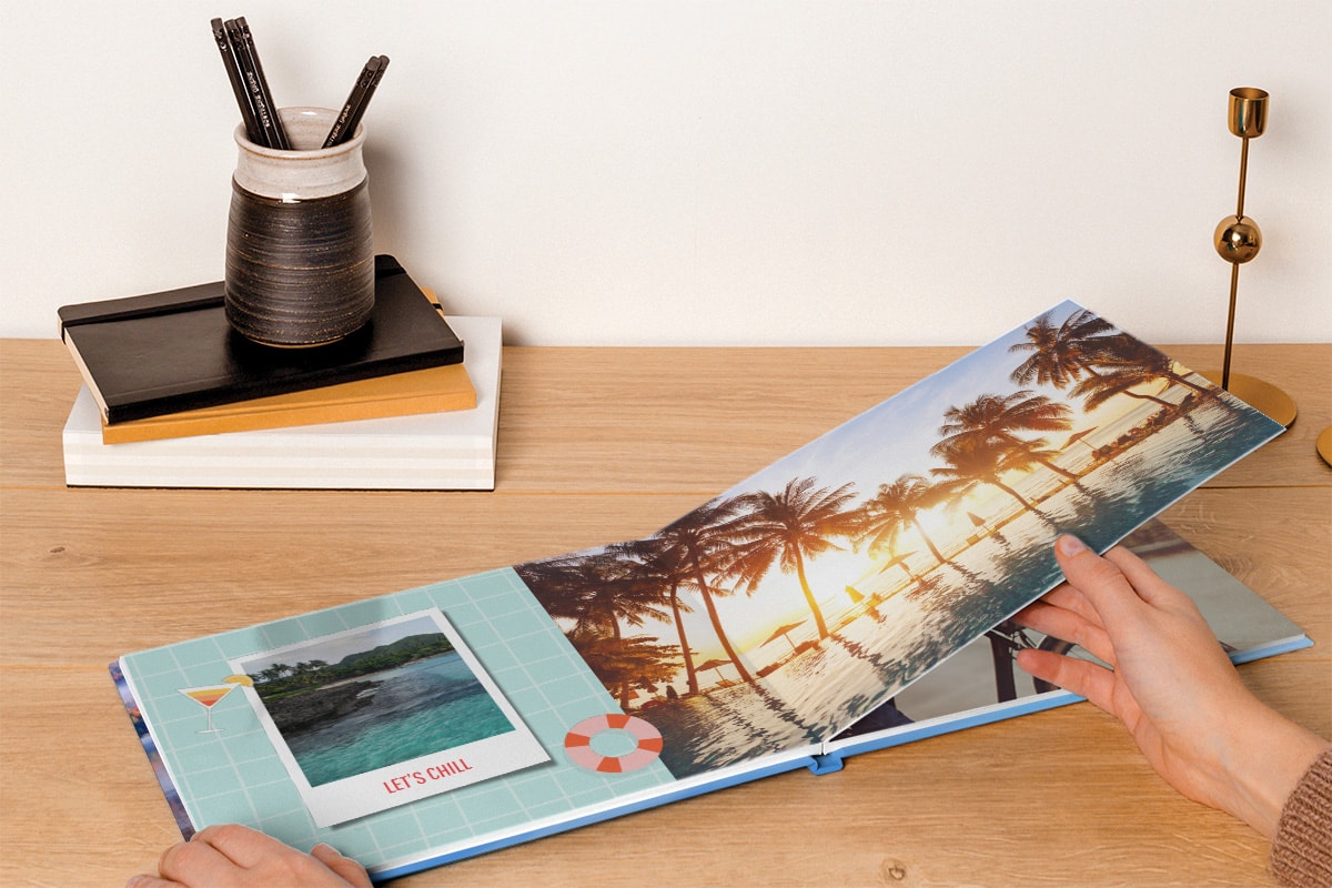 Die Hände einer Frau, die durch ein Fotobuch mit einer Layflat-Bindung blättern, das ein großes Foto von einem Pool bei Sonnenuntergang zeigt, welches sich über zwei Seiten erstreckt, mit einem kleineren Foto im Stil einer Sofortbildkamera, auf einem blaugrünen Hintergrund am Rand der linken Seite.