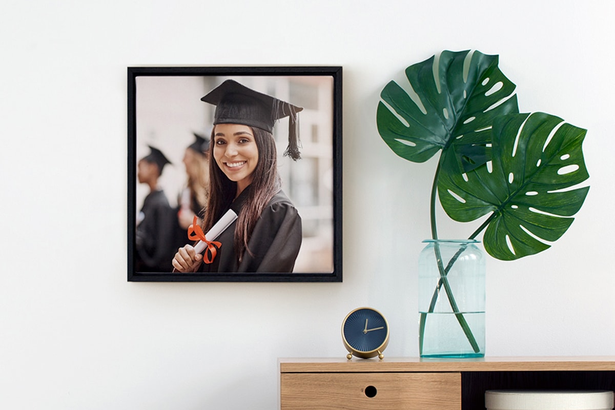 Ein Bild eines gerahmten Fotos auf Leinwand, mit einem Abschlussfoto von einem Mädchen. Es hängt an einer Wand, neben einem Beistelltisch mit einer Vase.