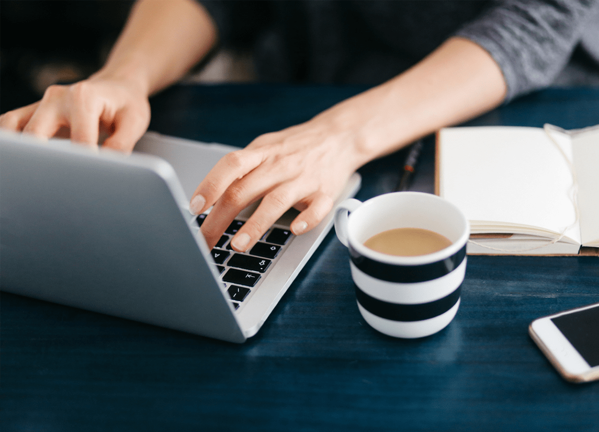 Zwei Hände, die auf der Tastatur eines Laptops tippen. Die Person sitzt an einem Schreibtisch mit einer Tasse Kaffee, einem Notizbuch und einem Telefon neben dem Laptop.