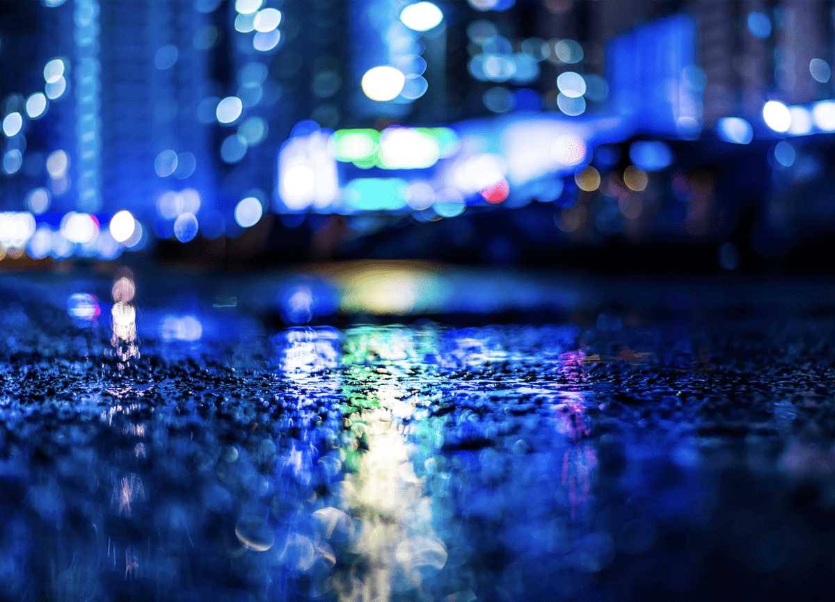 Ein Foto, das in einer regnerischen Nacht sehr nah am Boden gemacht wurde. Im Hintergrund sieht man helle Lichter, die verschwommen sind.