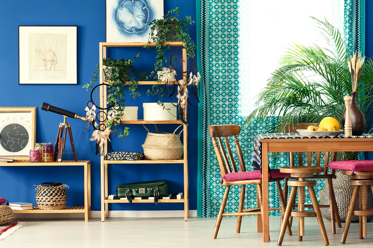 Ein Foto eines Zimmers mit einer royalblauen Wand, Regalen aus naturbelassenem Kiefernholz, blattreichen Zimmerpflanzen auf den Regalen und einem Tisch, mit von der Natur inspirierten eingerahmten Kunstwerken an der Wand.