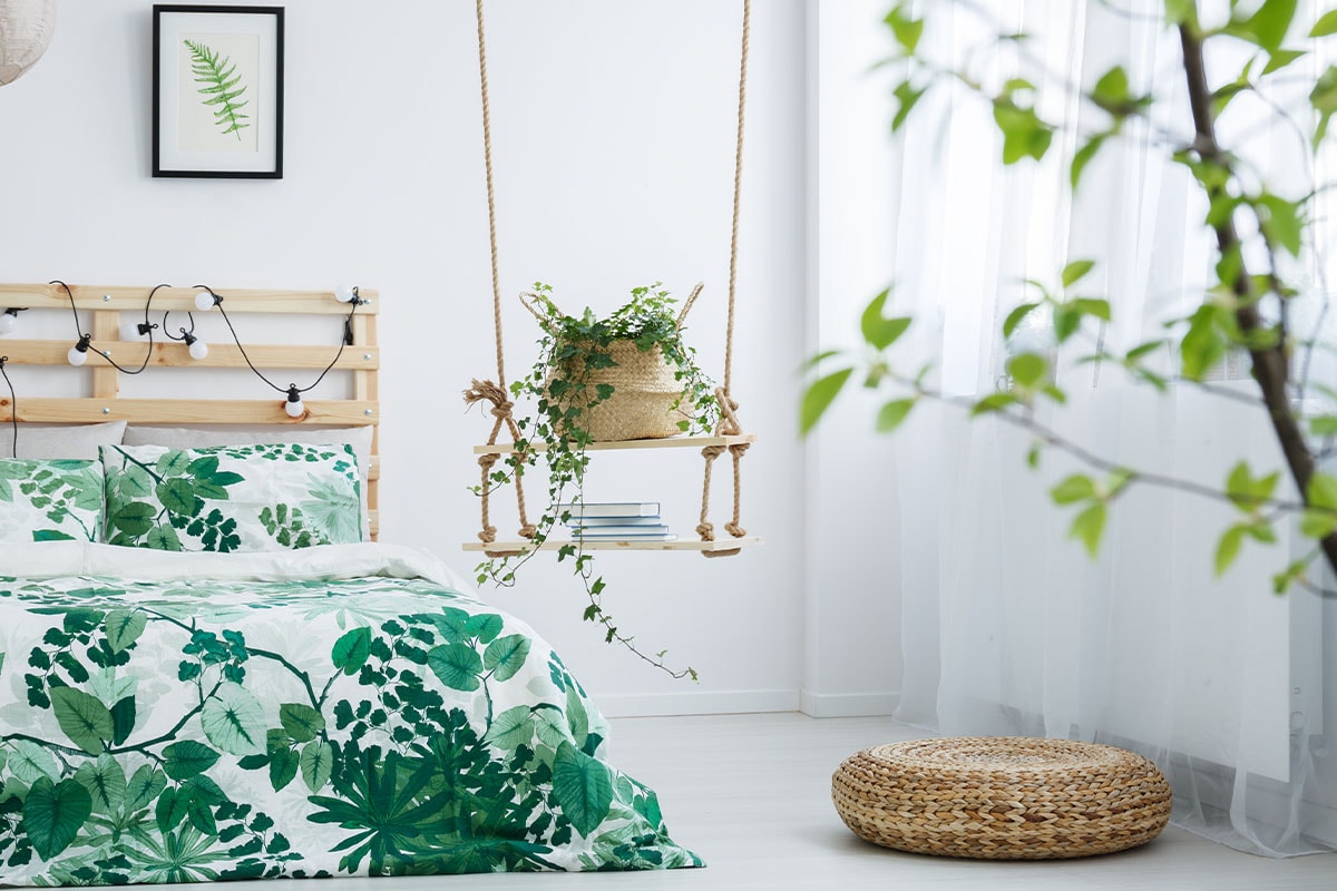 Ein helles Schlafzimmer mit einem Bett aus naturbelassenen Kiefernholz, darauf Bettwäsche bedruckt mit einem Blättermuster, von der Decke hängende Seilregale mit darin Zimmerpflanzen und ein Stapel Bücher.