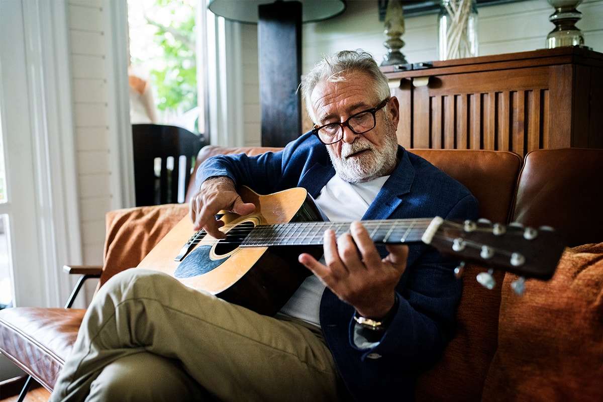Ein Foto eines älteren Mannes, der auf einer Couch sitzt und eine akustische Gitarre spielt.