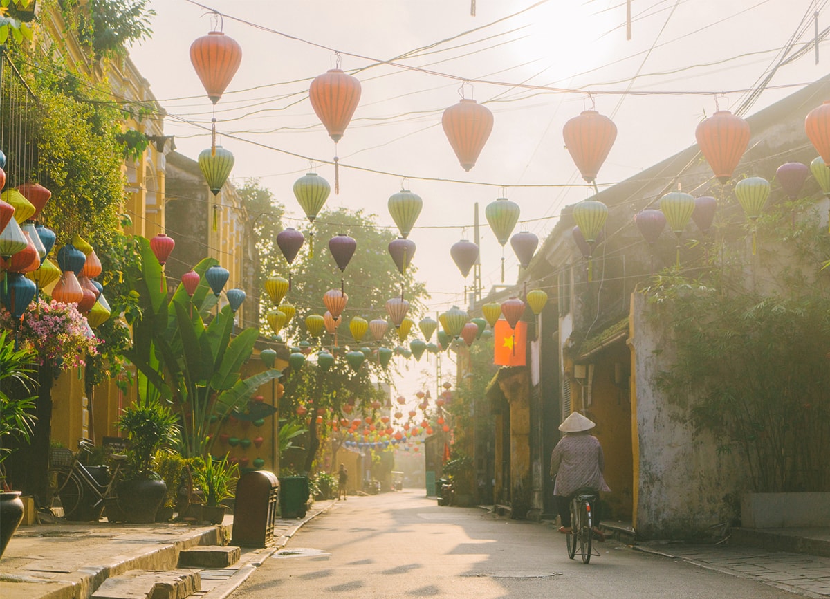 Frau, die auf einer Straße an einem trüben, sonnigen Tag Rad fährt. Über der Straße hängen bunte Papierlampions.
