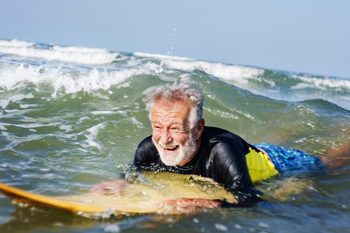 Ein älterer Mann lächelnd auf einem Surfboard am Meer.