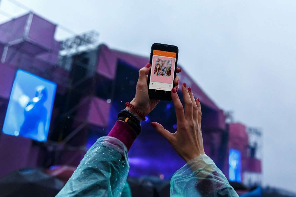 A photo of a woman holding up her phone in front of a stage at a festival, with a coloured retro print on the screen.