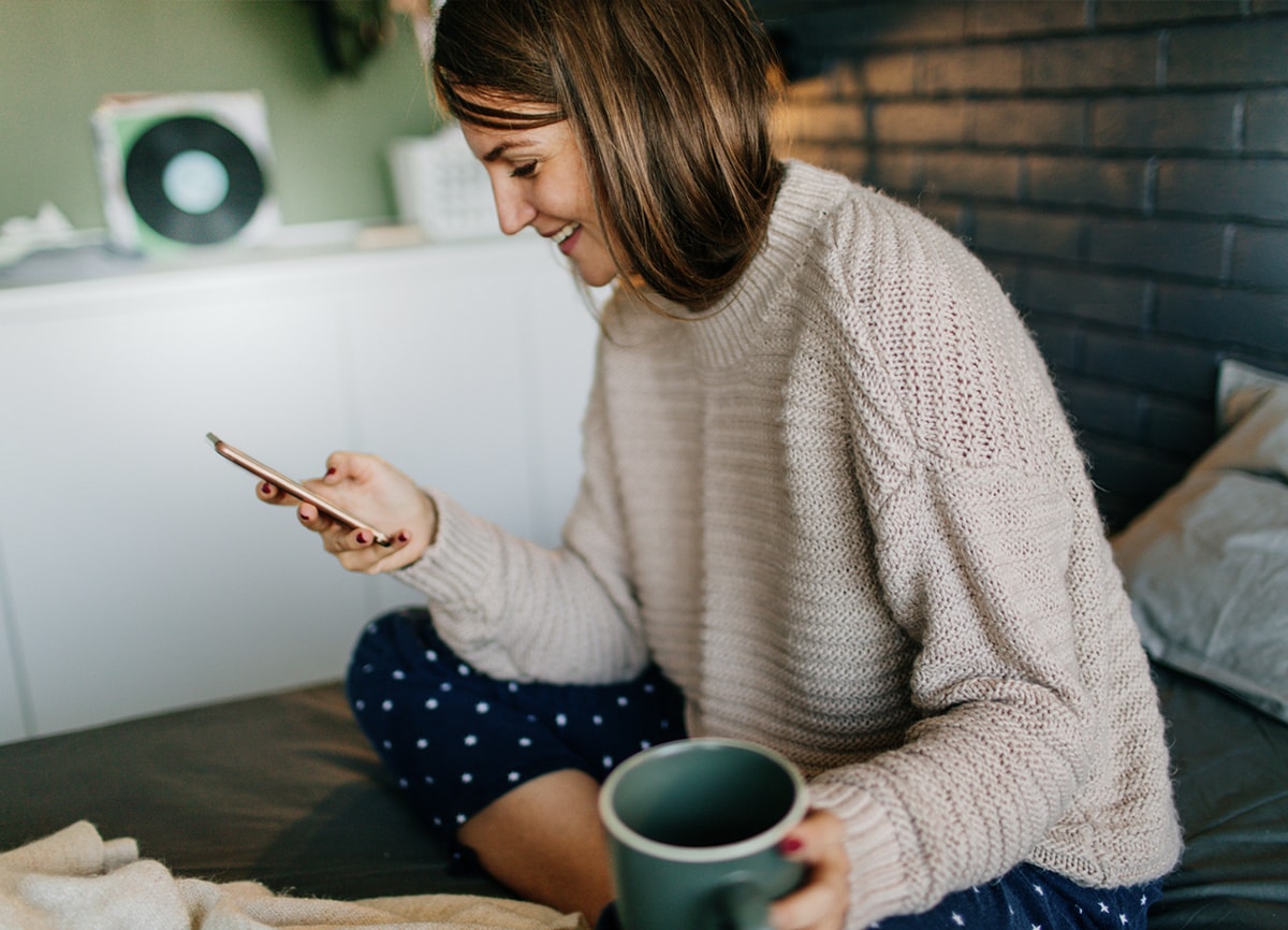 Een vrouw die zittend op bed op haar smartphone kijkt met een kop koffie in haar andere hand. 