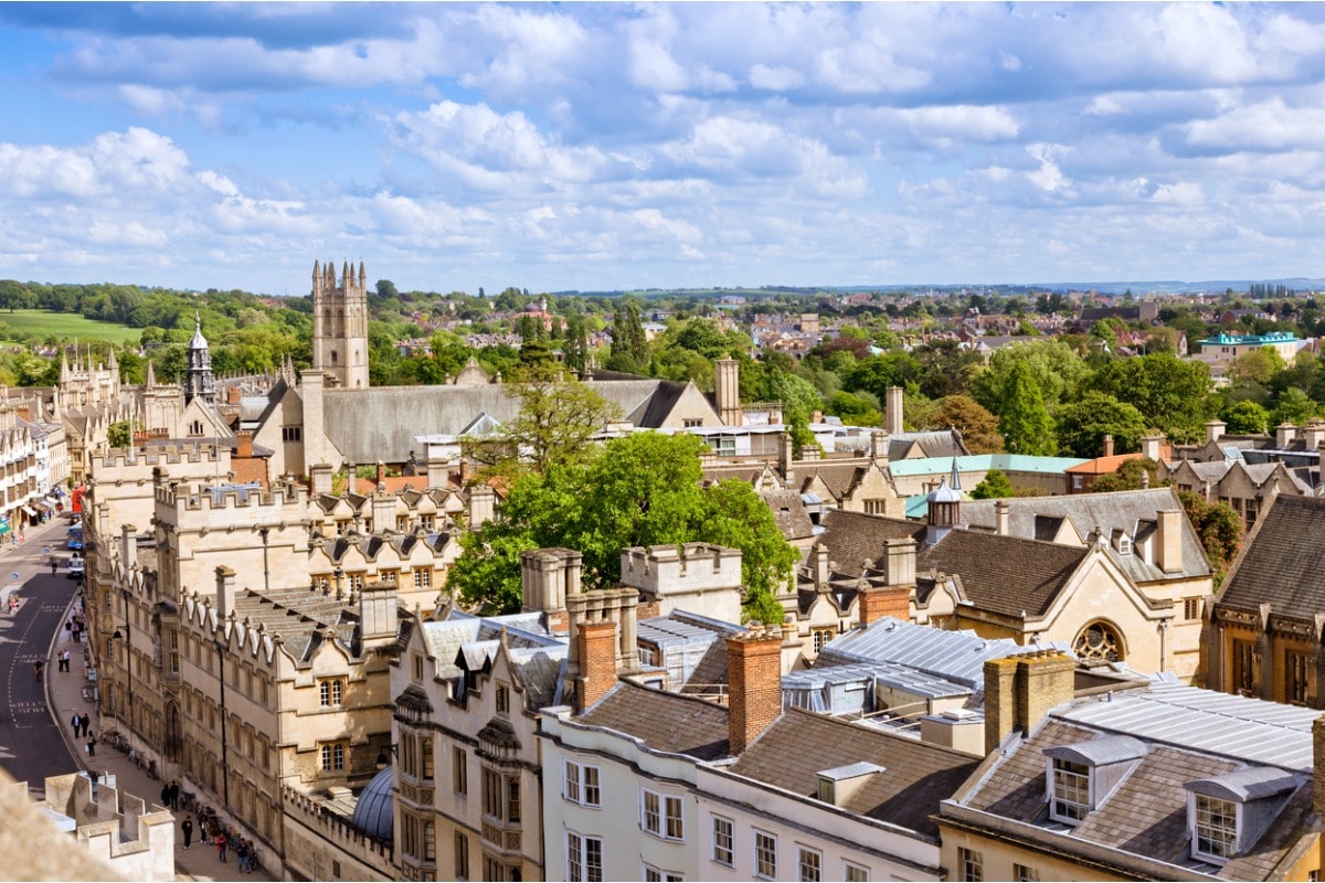 Een foto van de daken van Oxford en de klokkentoren van de kathedraal van de stad, met bomen en heuvels in de verte.