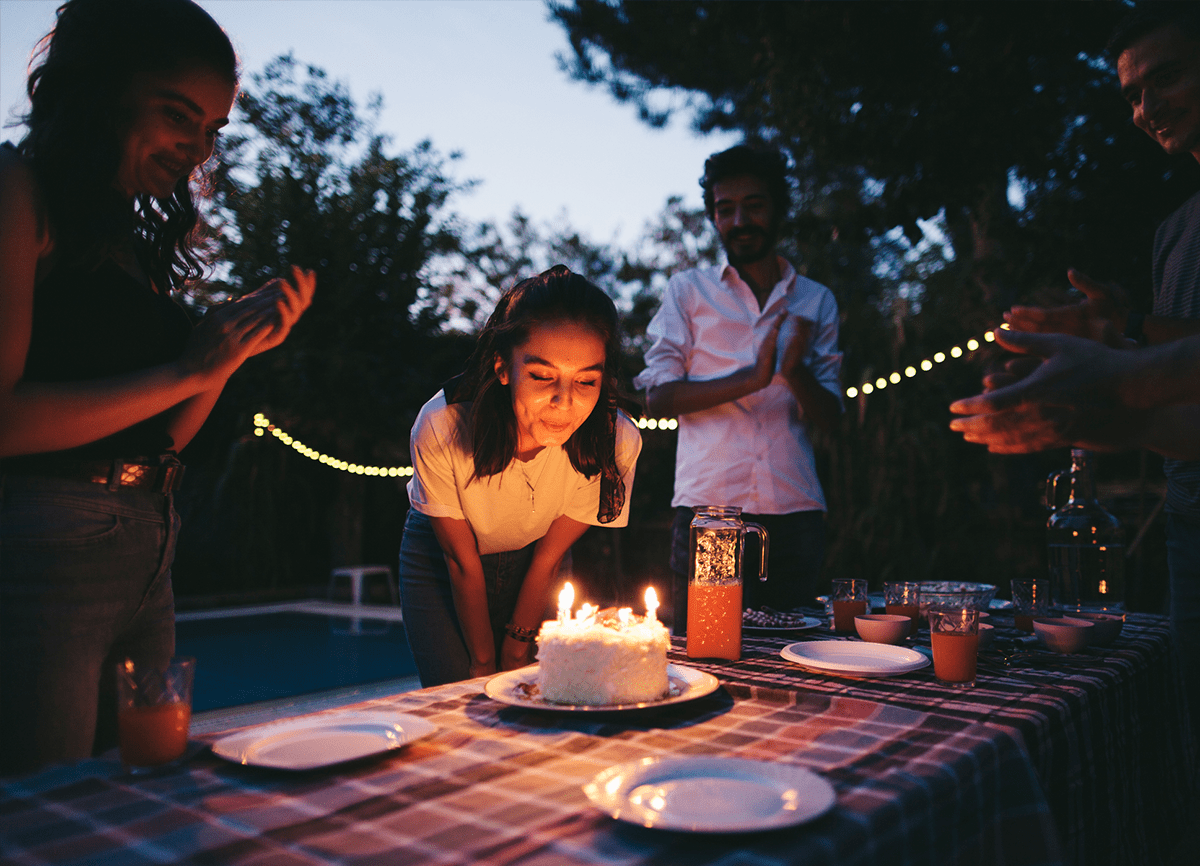 Een meisje dat kaarsjes op een verjaardagstaart uitblaast op een feestje buiten, 's avonds.