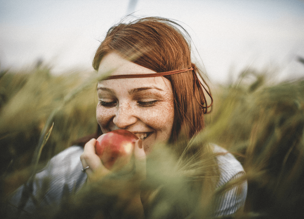 Een meisje dat een appel eet in een veld met hoog gras. Het gras op de voorgrond en achtergrond is ietwat onscherp, maar er is scherpgesteld op het meisje.