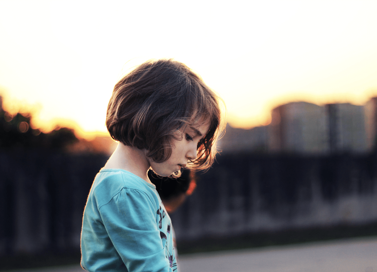 Een klein meisje dat buiten staat bij zonsondergang. De gebouwen op de achtergrond zijn wazig, maar het meisje is scherpgesteld.