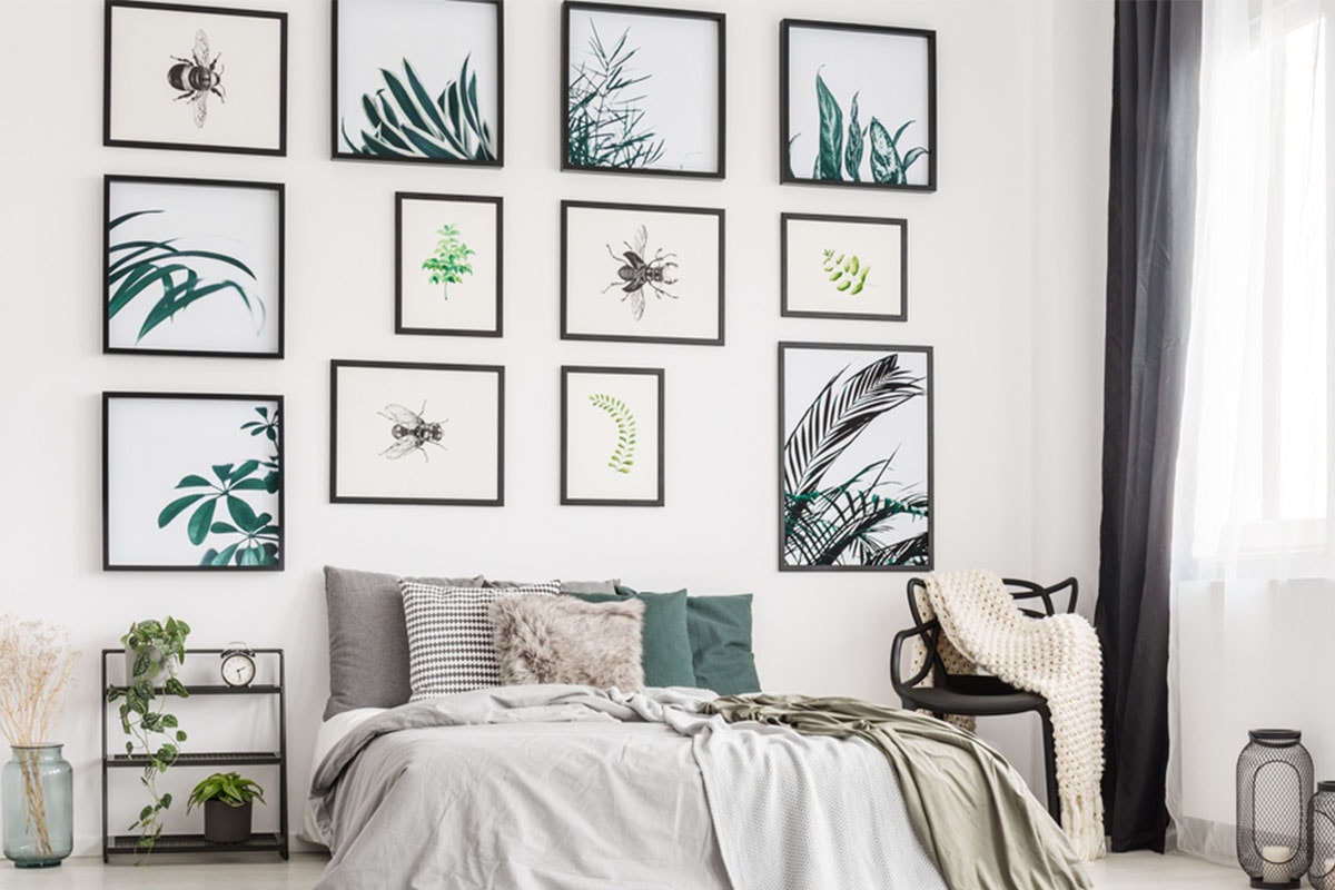 Een slaapkamer met lichtgrijze en groene meubelen, witte muren en een grote collectie afdrukken van verschillende groottes boven het bed, met illustraties van bladeren en insecten.