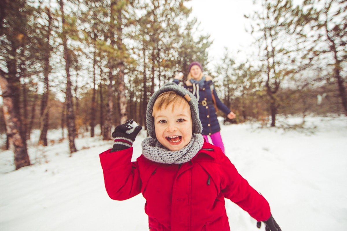 Foto van een kleine jongen in de sneeuw, klaar om een sneeuwbal naar de camera te gooien, met een vrouw op de achtergrond die ook een sneeuwbal gaat gooien.