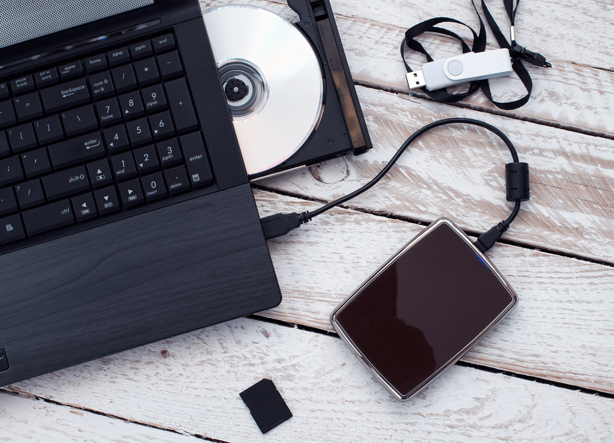 Een zwarte laptop met een DVD in een open CD-drive. Het staat op een wit houten tafelblad met een externe harde schijf, een USB-stick erboven en een geheugenkaart eronder. 
