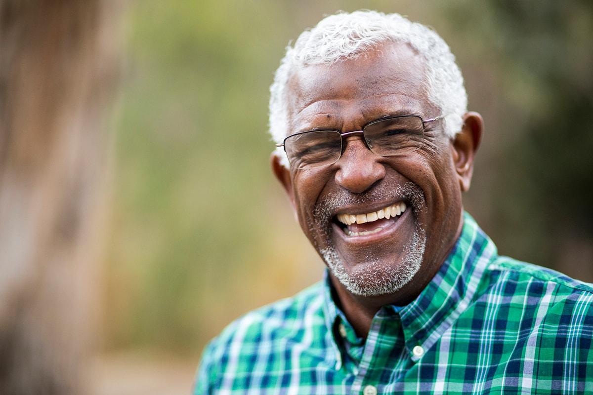 Een portretfoto van een oude lachende man. Op de achtergrond zie je wazige bomen, maar de man zelf is scherp.