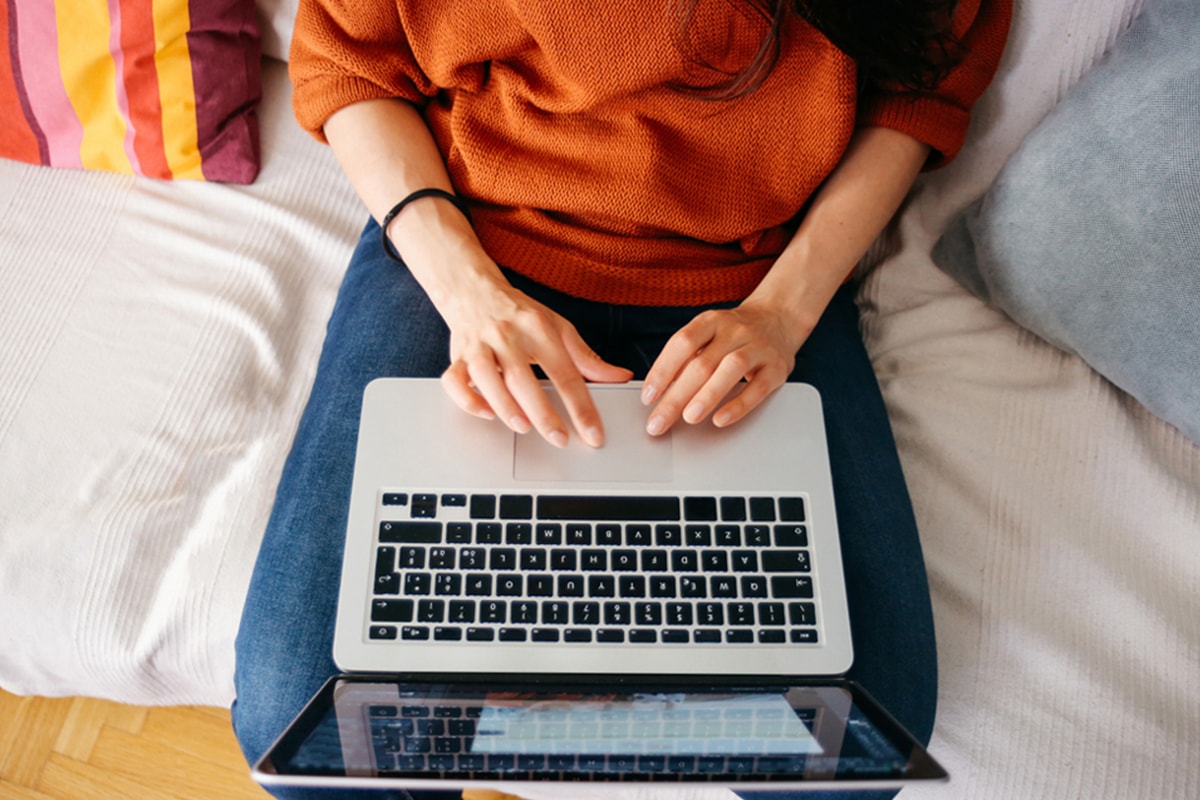 Een jonge vrouw op een grijze zetel in een witte woonkamer met een laptop op haar knie. Op de zwarte balk onderaan de pagina zie je een winkelwagen.