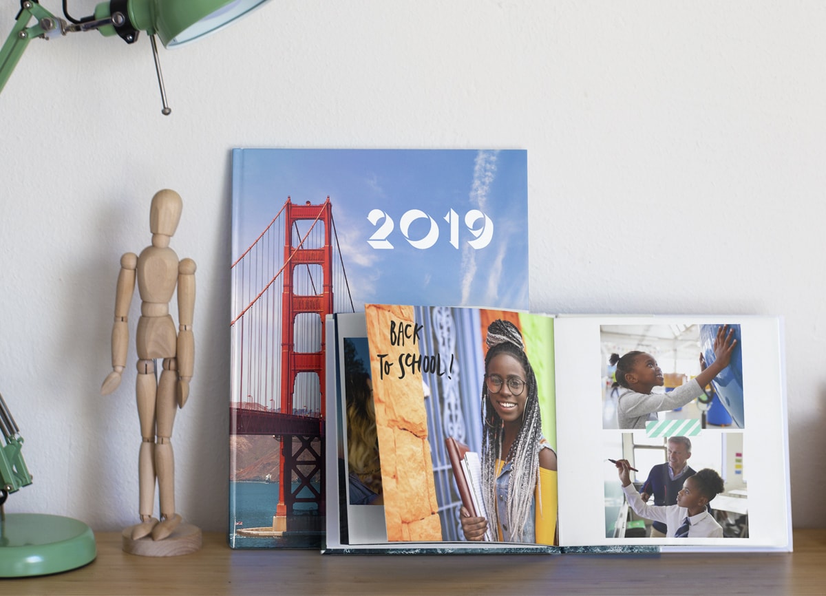 Två fotoböcker på en hylla. Fotoboken i bakgrunden visar en bild av San Francisco på omslaget, och den som är i förgrunden ligger öppen och visar bilder av en ung flicka under olika skolstadier: en bild av henne på högskolan, en i förskolan och en annan i grundskolan. 