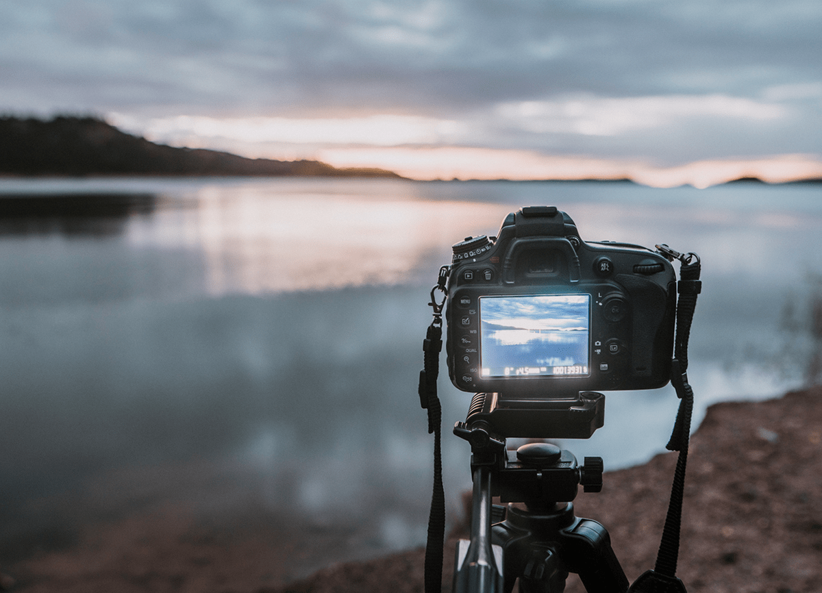 En DSLR-kamera på ett stativ riktad mot en sjö i svagt ljus. Kameran är i fokus, bakgrunden är oskarp.
