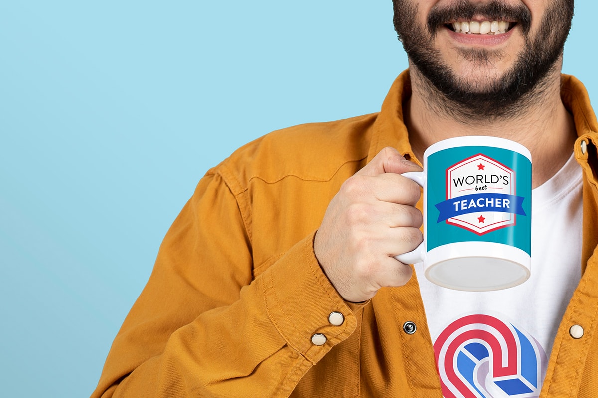A man in a mustard-coloured shirt, against a blue background, holding a customised mug with ‘world’s best teacher’ on.