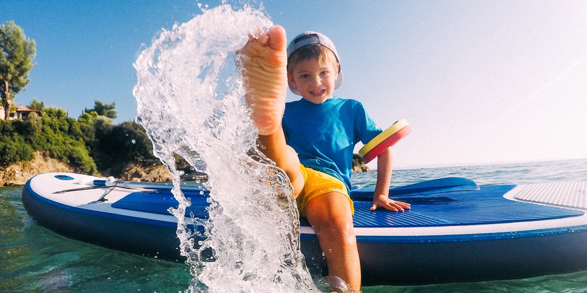 En pojke står på en SUP-bräda i havet och stänker vatten mot kameran.