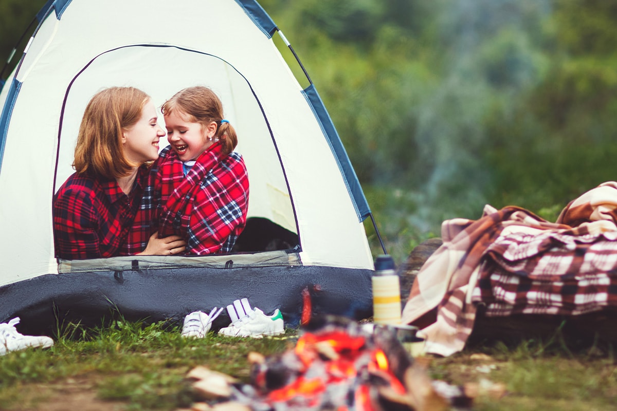 En mamma med dotter ler i ett tält framför en lägereld