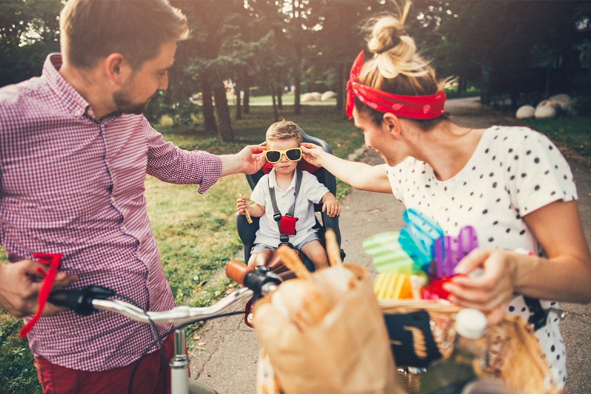 Et ungt par på sykkeltur i parken, som setter solbriller på sønnen sin som sitter i et sykkelsete.